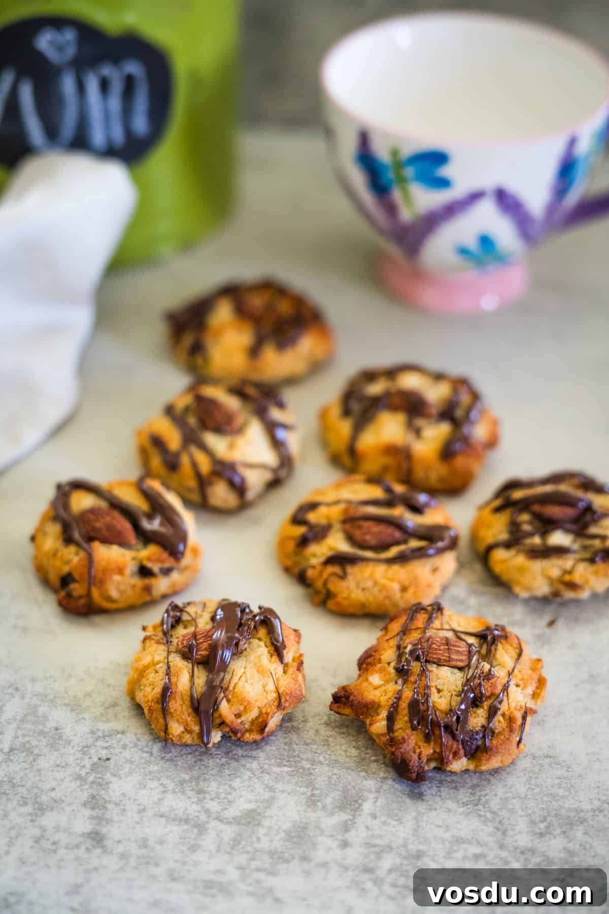 Keto Almond Joy Cookies Recipe — Low-Carb Coconut Chocolate Cookies 10 Keto almond joy cookies topped with almonds and drizzled with chocolate are arranged on a table. A tea cup and a jar sit in the background.