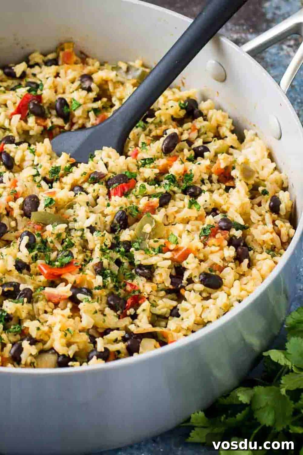 Side close up shot of beans and rice in a large saucepan with a black spatula inside all on a blue surface with fresh herbs in the background.