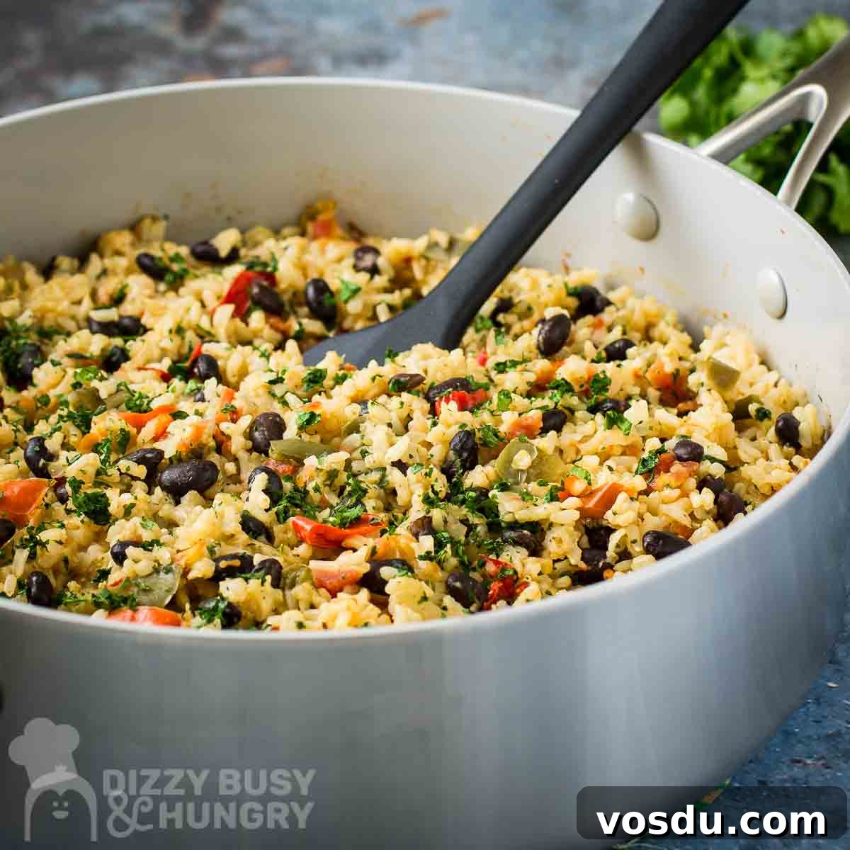 Side shot of beans and rice in a large saucepan with a black spatula inside all on a blue surface with fresh herbs in the background.
