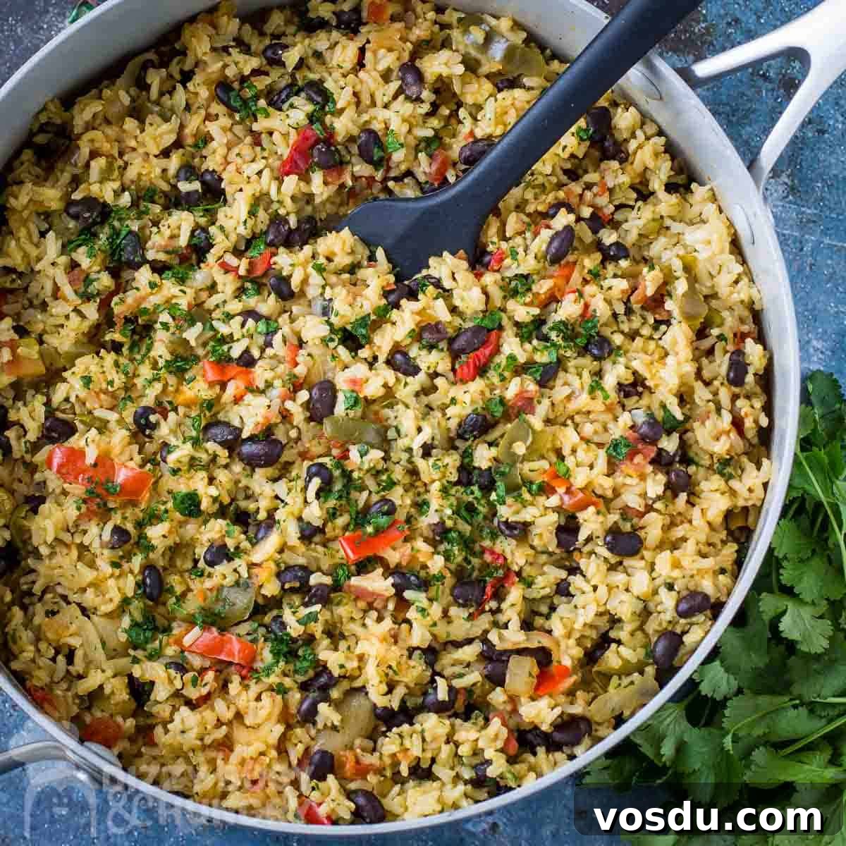 Overhead shot of beans and rice in a large saucepan with a black spatula inside all on a blue surface with fresh herbs in the background.