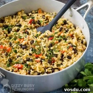 Side angled shot of beans and rice in a large saucepan with a black spatula inside all on a blue surface with fresh herbs in the background.