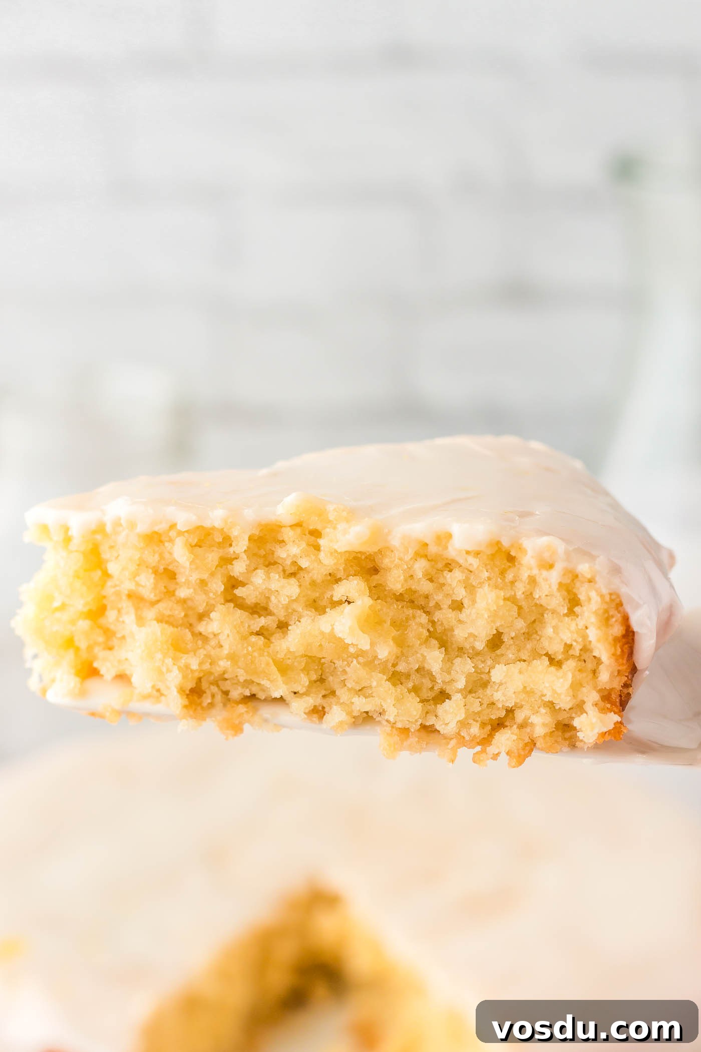 A slice of vegan olive oil cake topped with powdered sugar glaze held on a spatula over the whole cake below it.