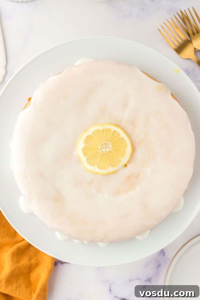 Overhead view of an olive oil cake topped with a powdered sugar icing on a cake stand. There's a slice of lemon decorating the middle of the cake.
