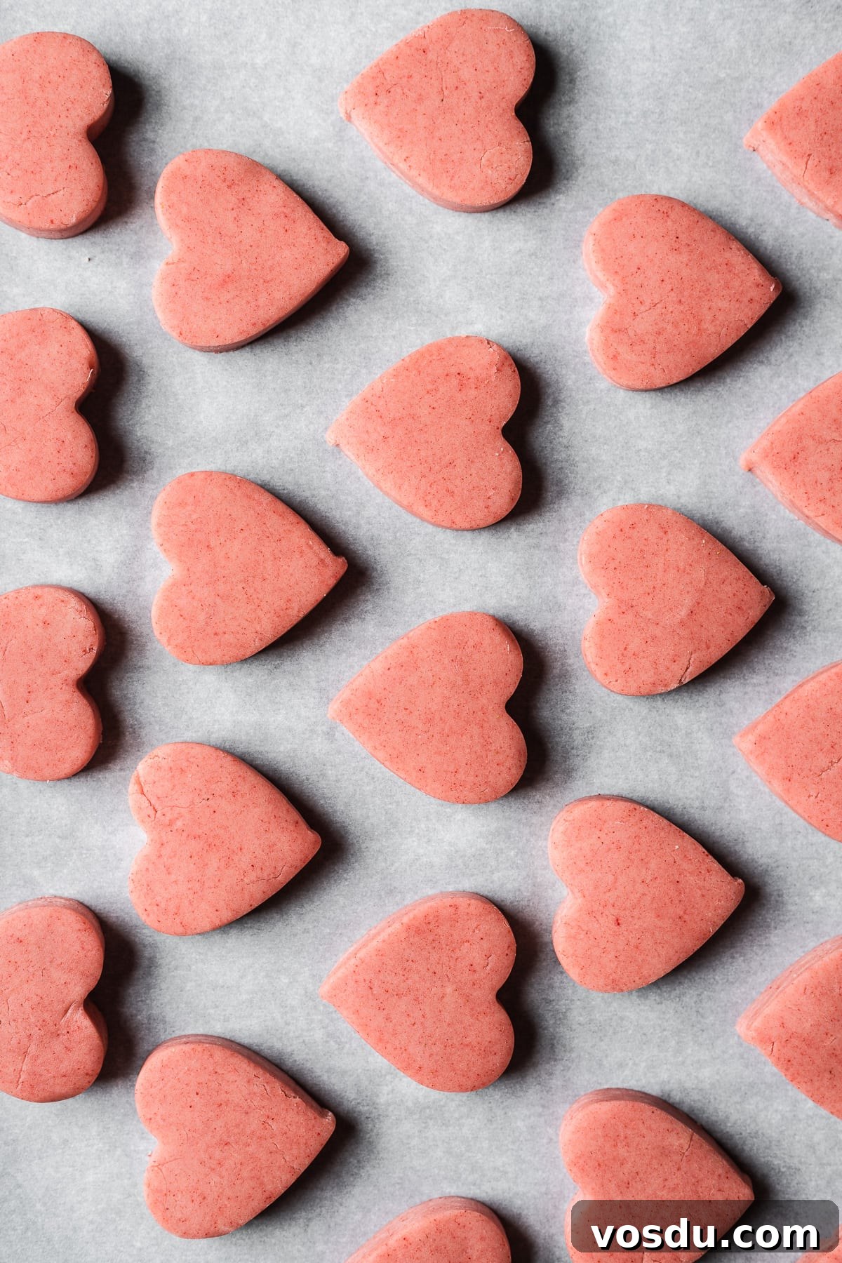 Strawberry Shortbread Cookies Recipe — Simple Homemade Treat 12 Rows of unbaked pink heart shaped cookies on white parchment paper.