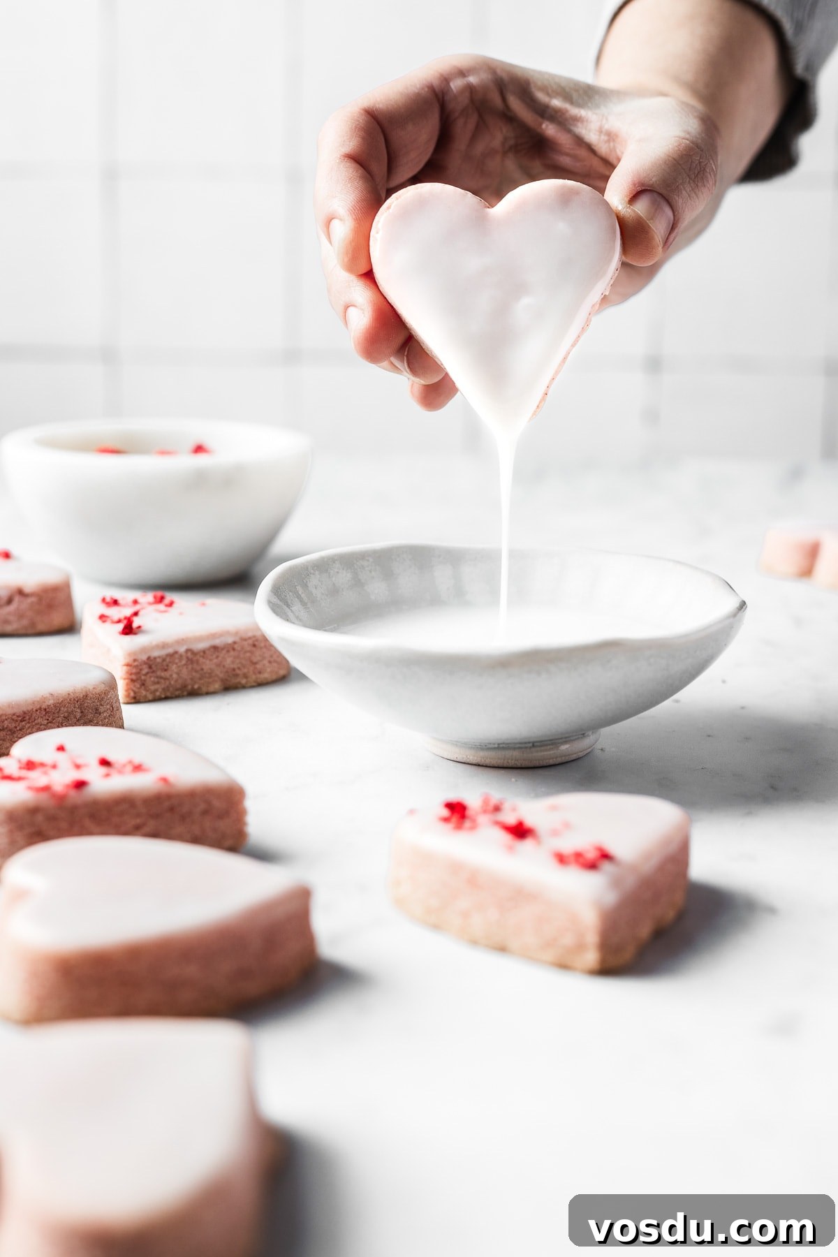 Strawberry Shortbread Cookies Recipe — Simple Homemade Treat 13 A hand dipping a pink shortbread cookie into a bowl of glaze.
