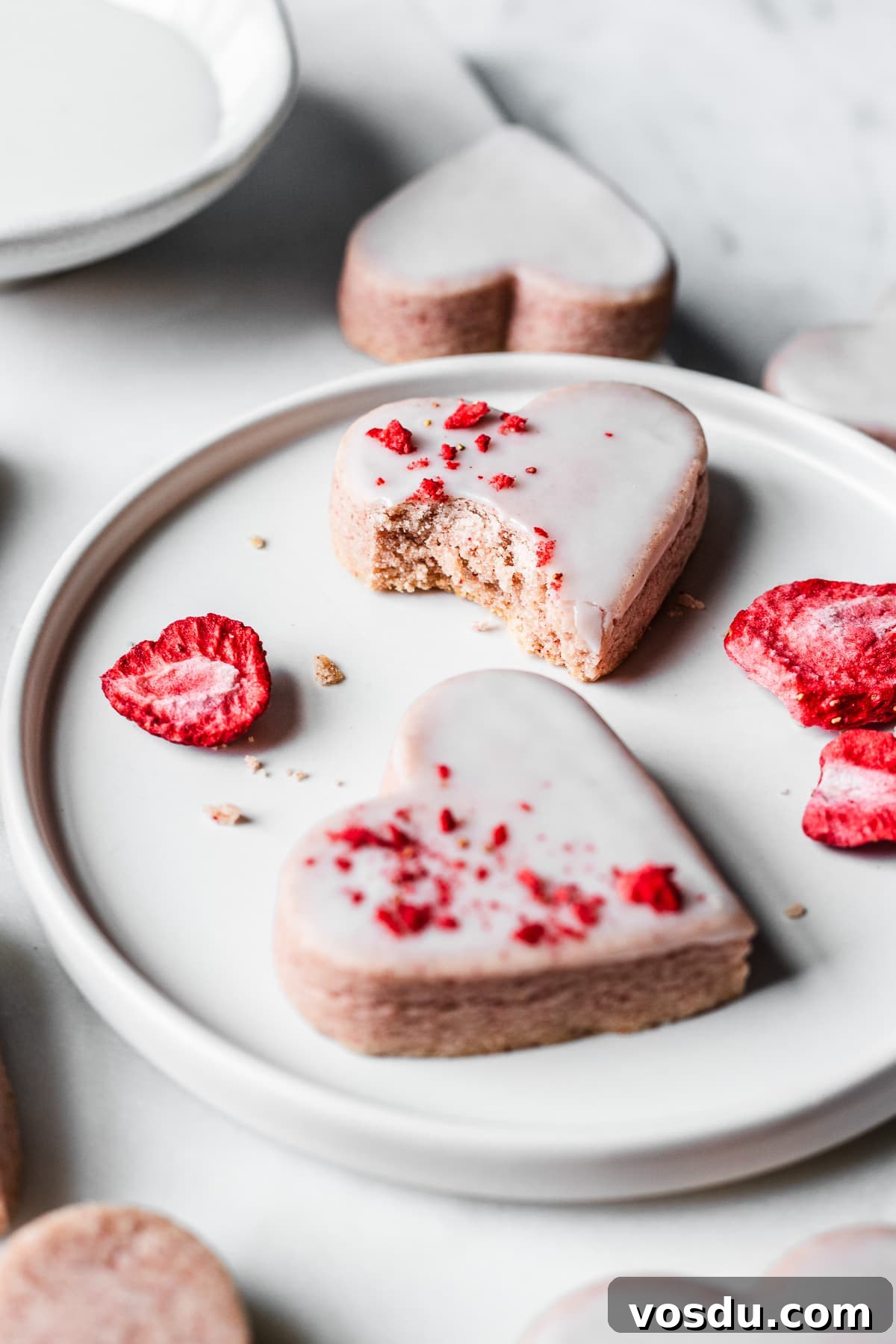 Strawberry Shortbread Cookies Recipe — Simple Homemade Treat 15 Two glazed heart shaped cookies on a plate. One has a bite out of it.