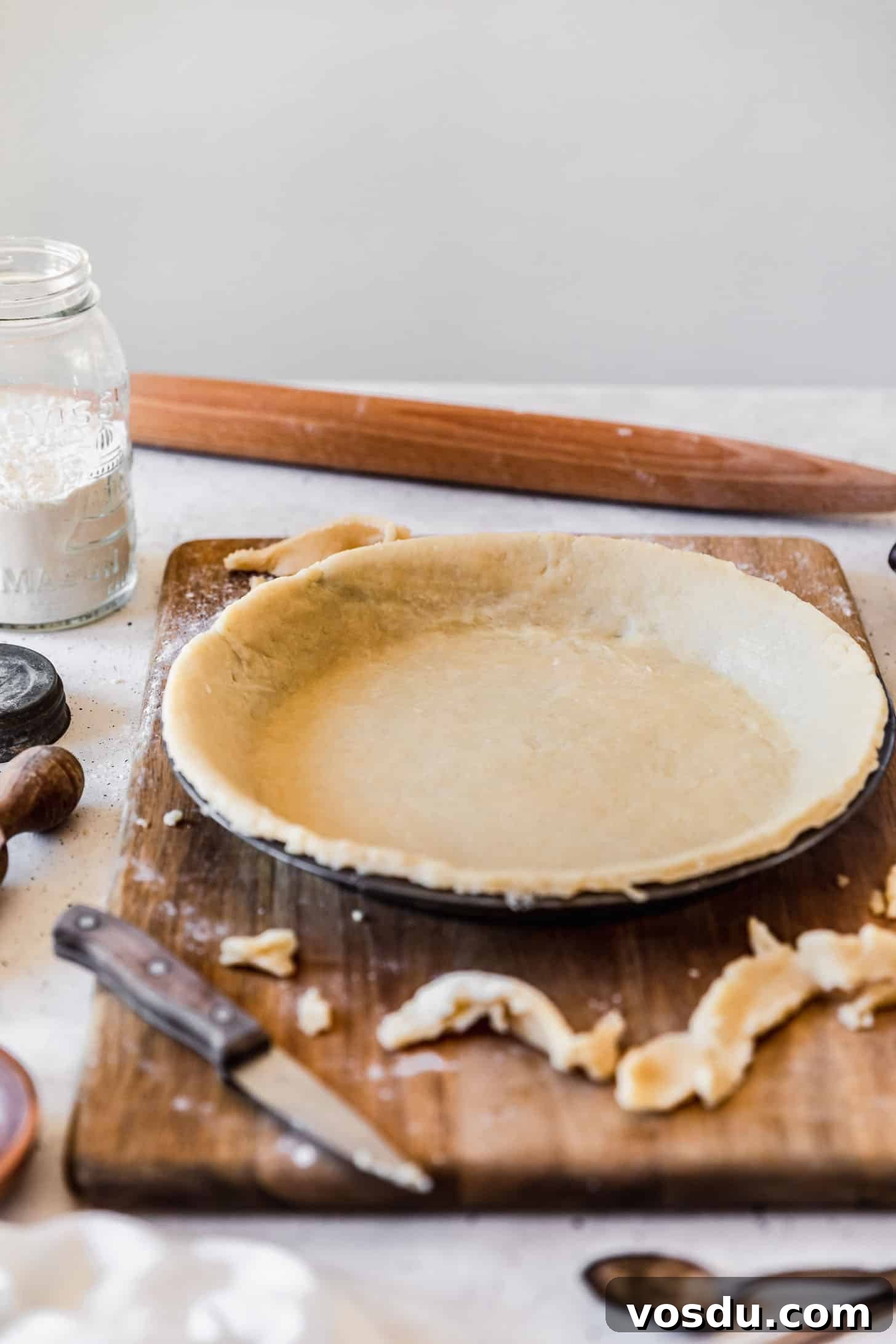 The Ultimate Pie Crust Recipe That Will Wow Your Guests 2 The best pie crust in a metal pie plate on a wood board next to pie tools and ingredients in front of a white wall.
