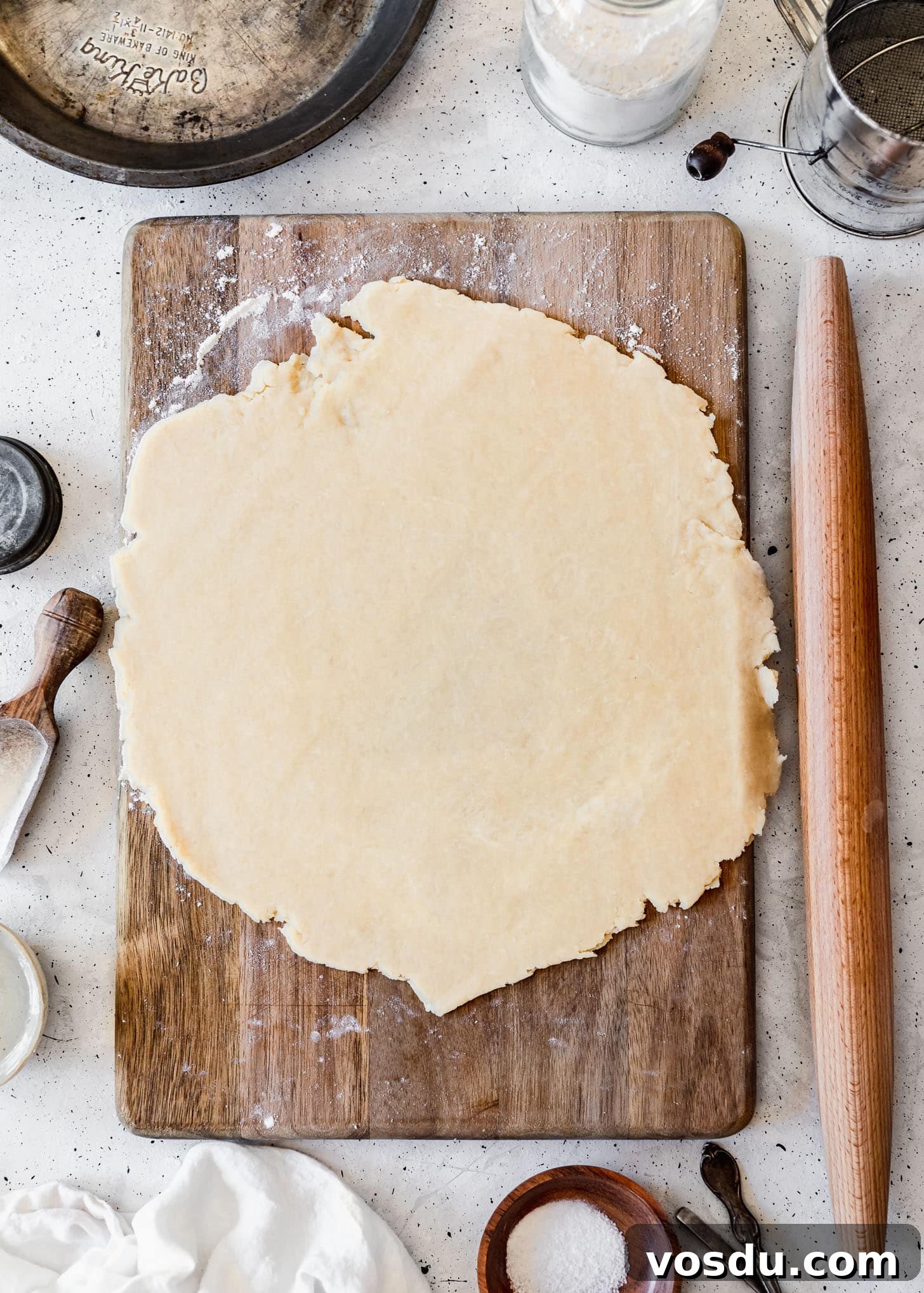 The Ultimate Pie Crust Recipe That Will Wow Your Guests 10 The best pie crust rolled out on a wood board next to baking tools and ingredients with a grey background.