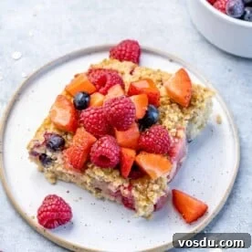 side shot of a slice of baked oatmeal on a white plate topped with berries and maple syrup