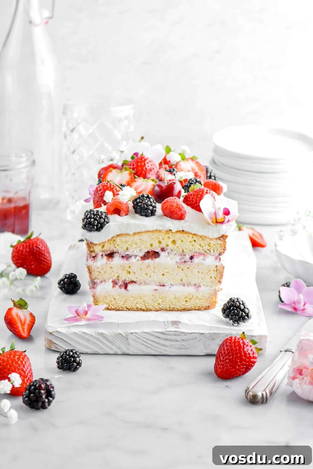 ice cream pound cake on white wood board with flowers and berries on top and surrounding cake, a stack of white plates, two empty glasses, and a jar of strawberry jam behind cake on marble surface.