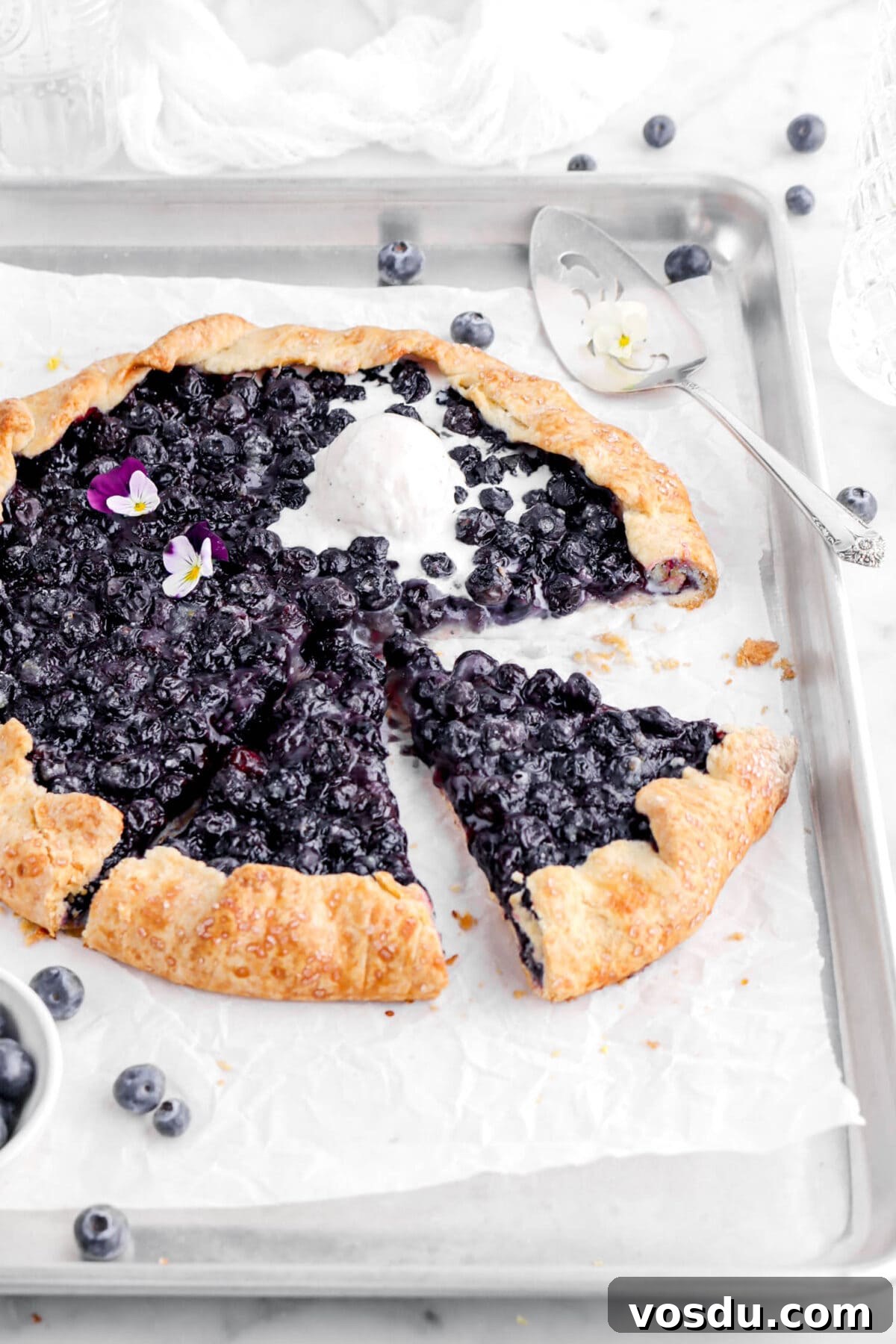 angled close up of sliced blueberry galette with melting scoop of ice cream on top, with cake knife and blueberries behind.
