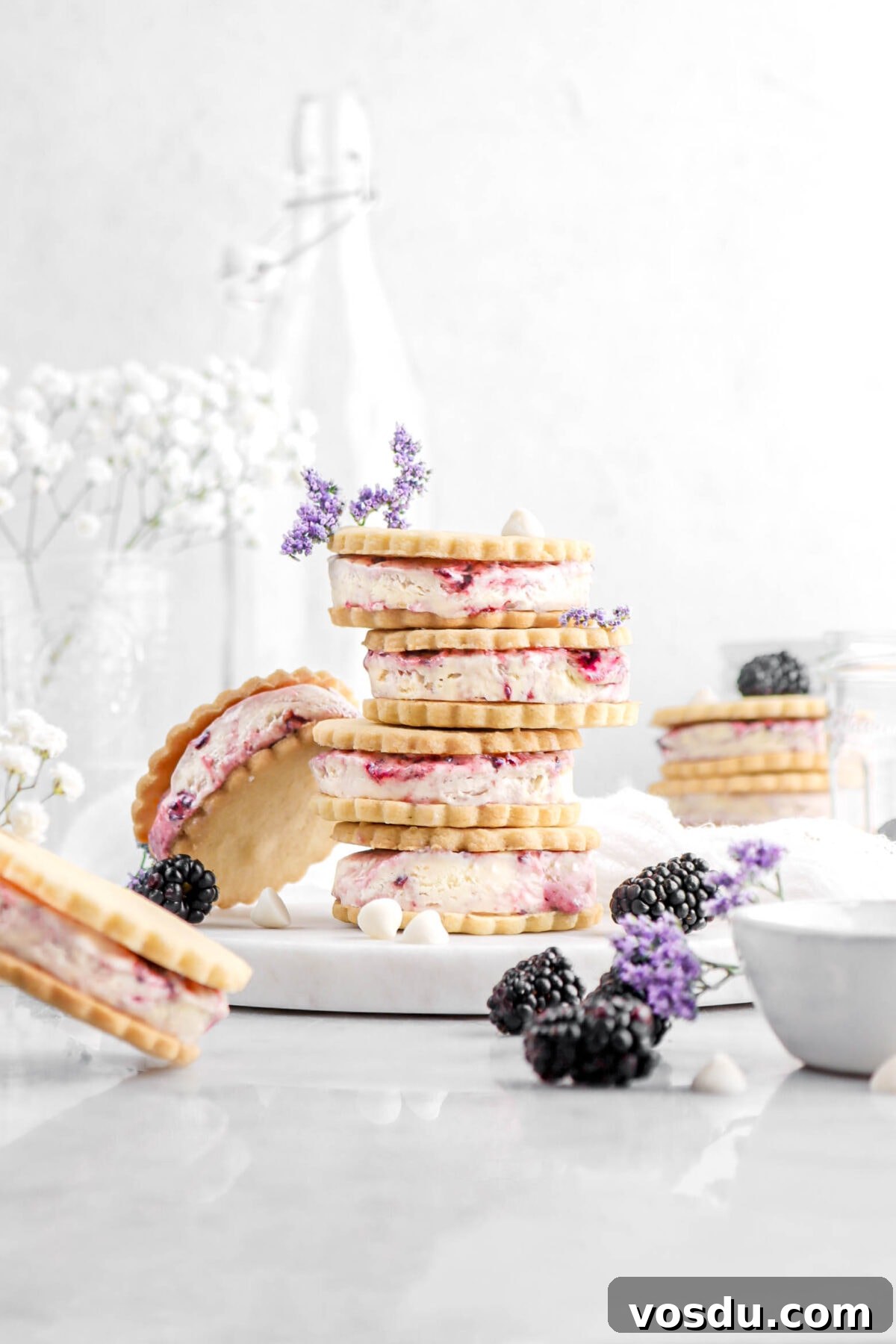 four stacked blackberry white chocolate ice cream sandwiches with fifth ice cream sandwich leaning against stack, with more ice cream sandwiches around on marble surface with blackberries, purple and white flowers, and white chocolate chips around.