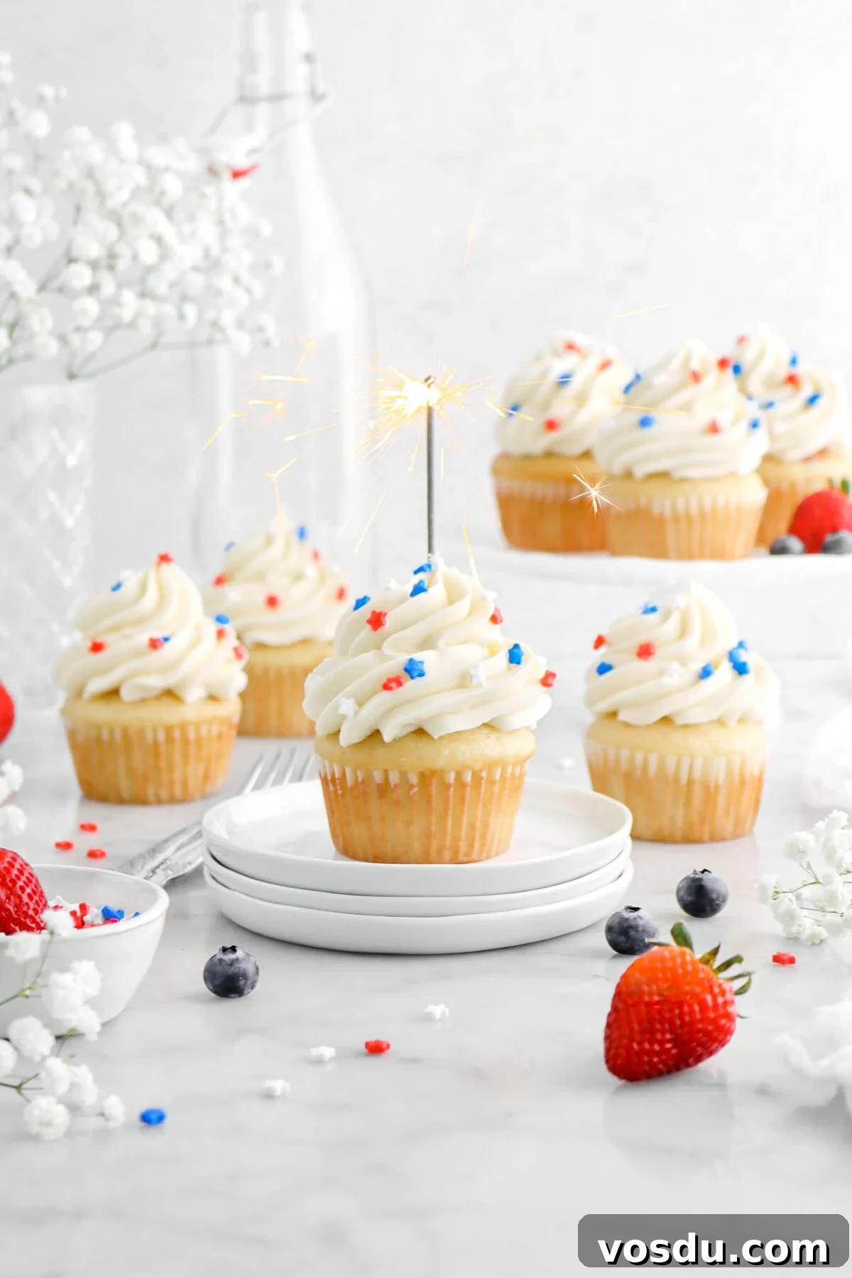 vanilla cupcake with frosting and red, white, and blue sprinkles on top with lit sparkler in cupcake with more cupcakes around, white flowers, and berries surrounding cupcake.