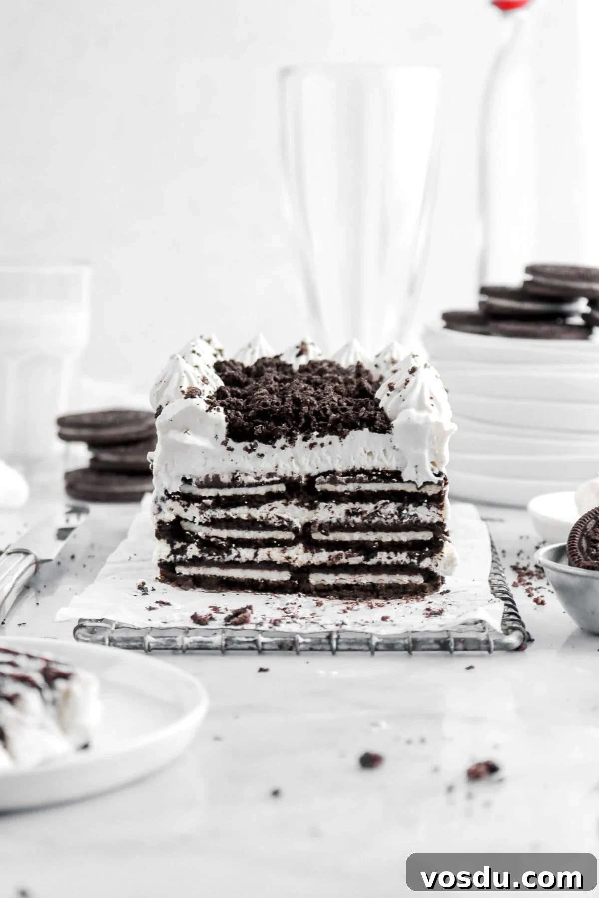 oreo icebox cake on wire tray with slice of cake on small white plate beside, with oreos around, a glass of milk behind, a stack of white plates, and two empty glasses.