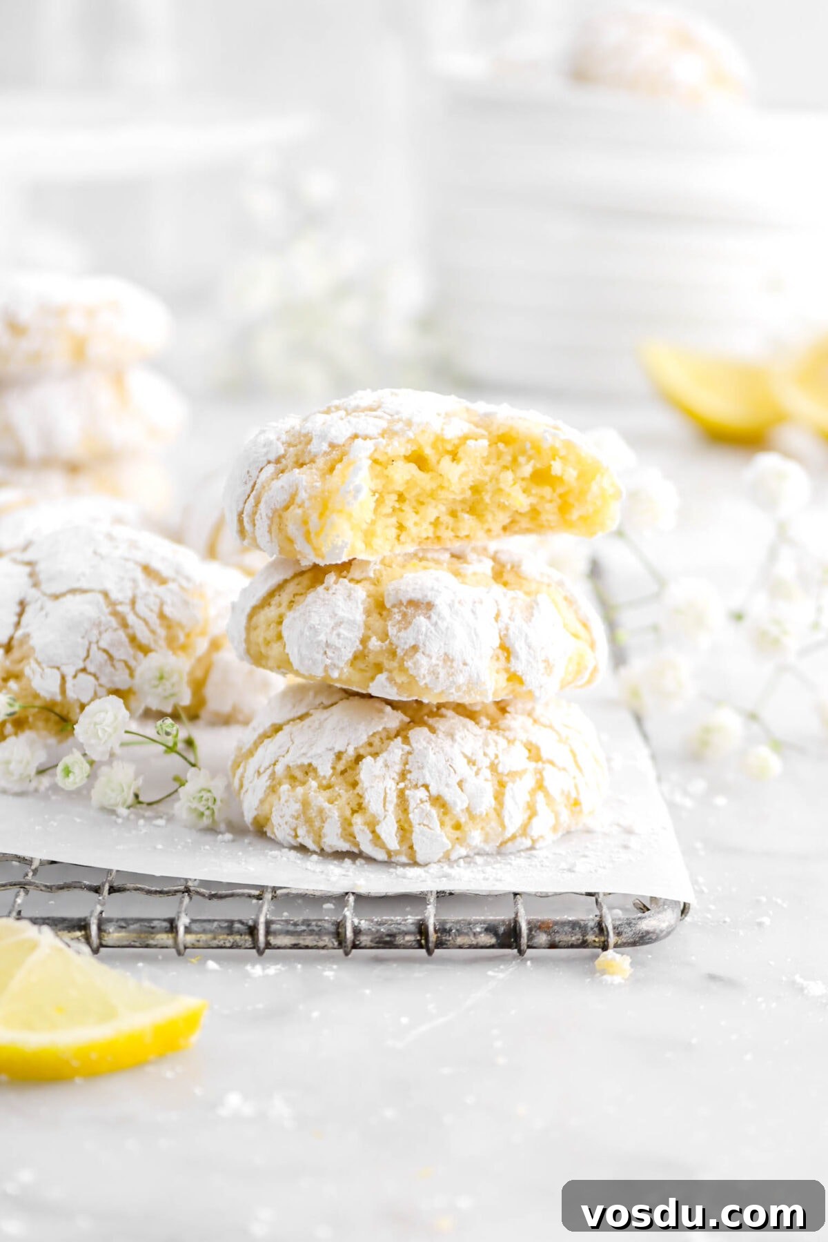 three stacked lemon crinkle cookies with bite taken out of the one on top with more cookies, flowers, and lemon slices behind