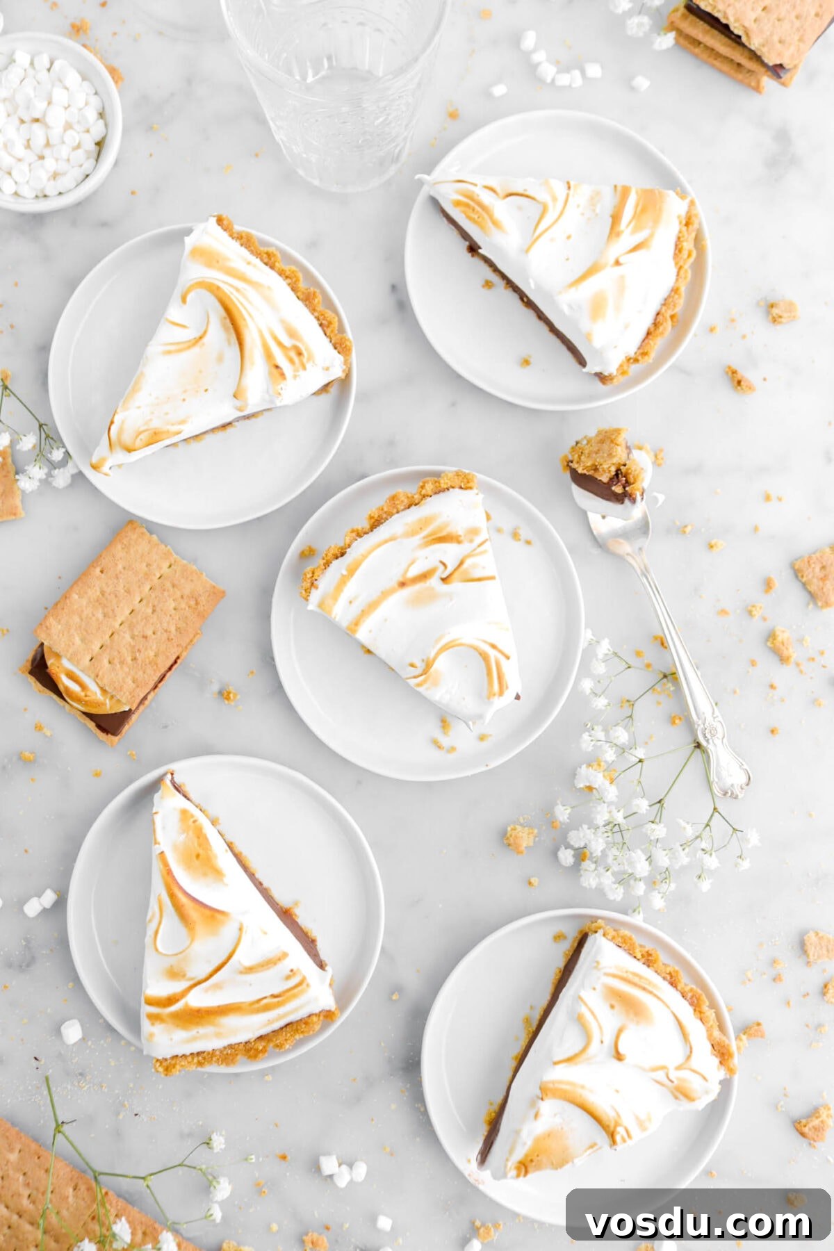 overhead shot of five slices of s'mores pie on marble surface with fork beside, white flowers, graham cracker pieces, white flowers, and mini marshmallows.