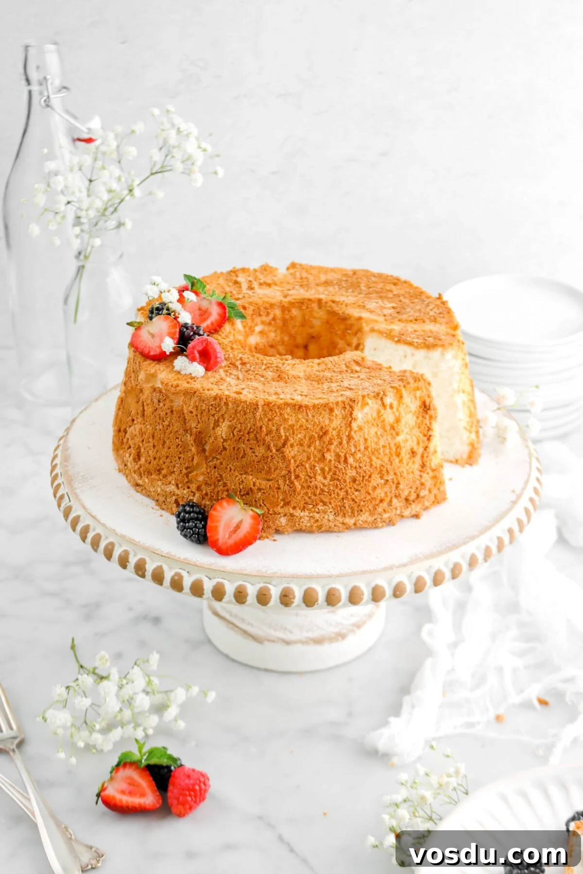 angel food cake with slice missing on white washed cake board with berries on top, fresh mind sprig, and white flowers with cheesecloth beside and forks.