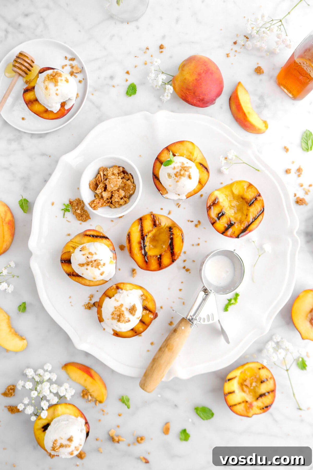 overhead shot of grilled peaches on round platter with ice cream scoop and bowl of crumble, ice cream toppiong three peaches, with mint leaves, more peaches, and white flowers around