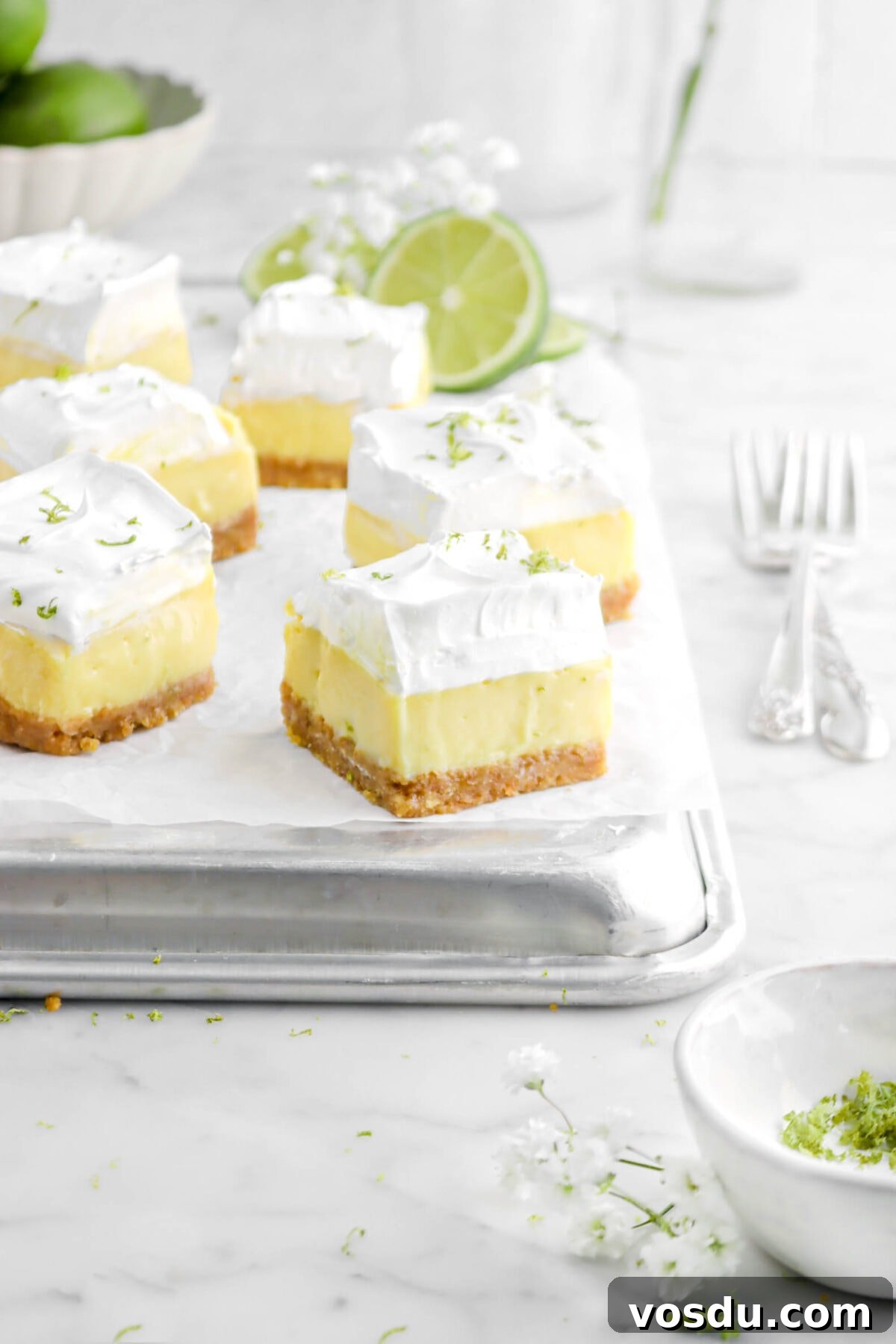 six key lime pie bars on sheet pan with sliced lime behind with white flowers, forks beside, and small white bowl of lime zest in front on marble surface