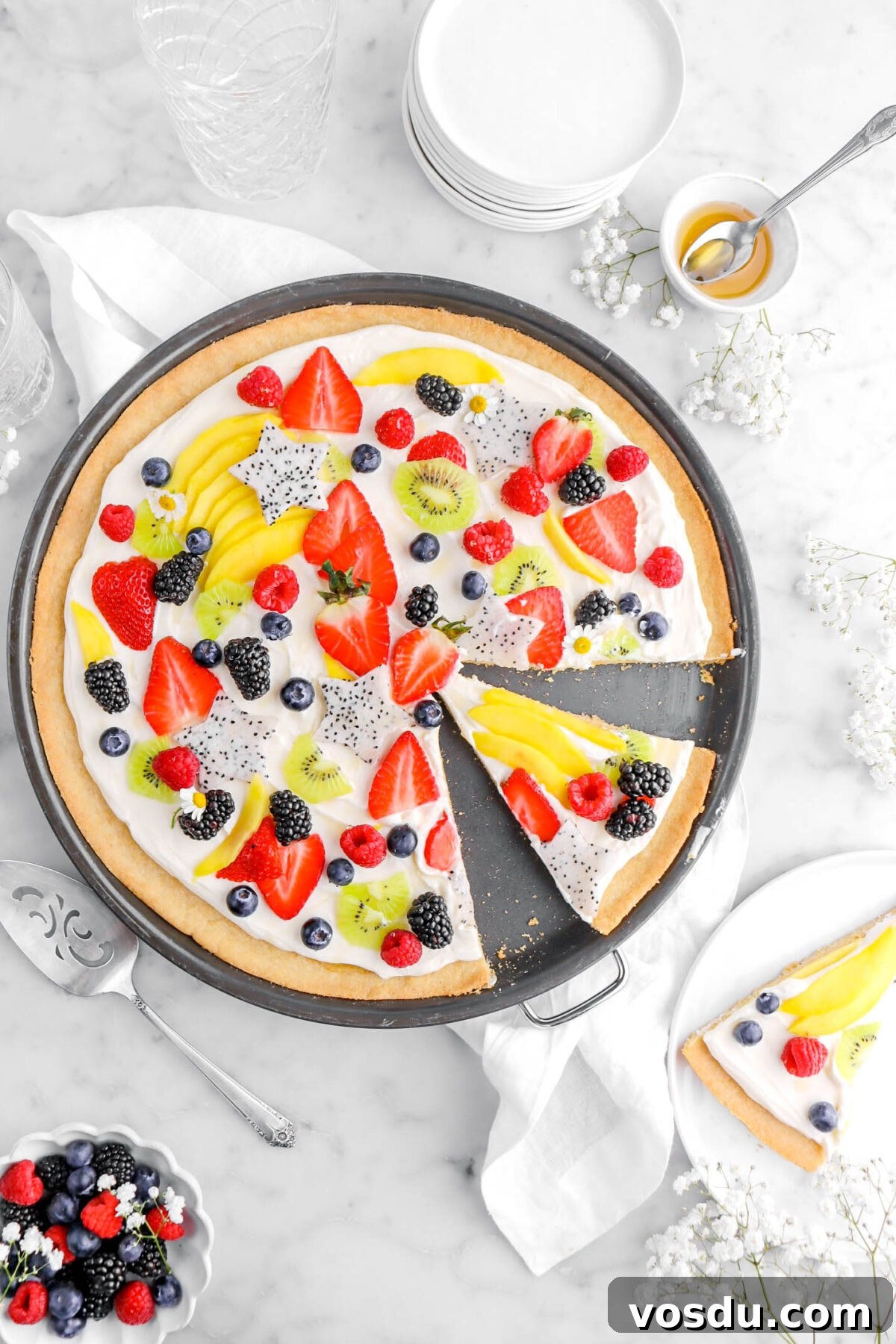 fruit pizza with slice cut into it in black pizza pan with white plate with another slice of pizza beside with bowl of berries and white flowers around.