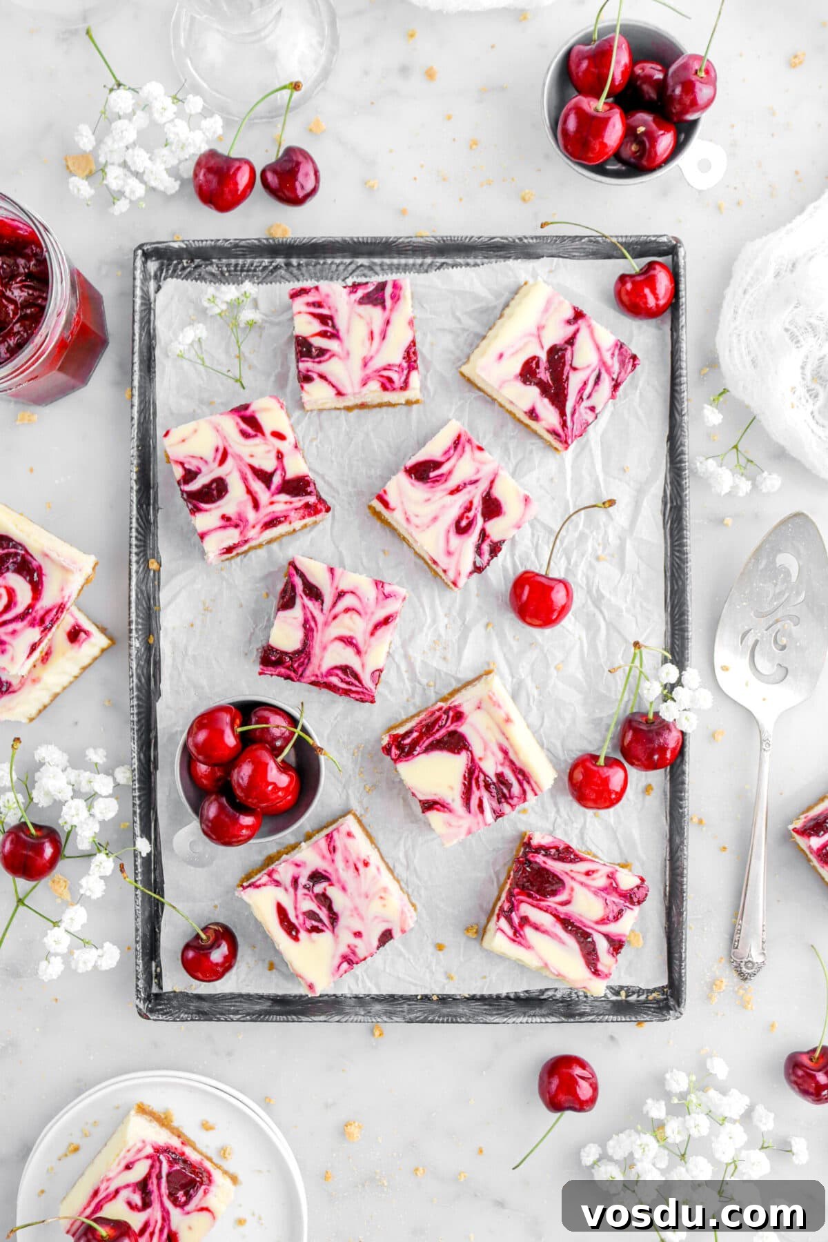 overhead shot of cherry swirled cheesecake bars on lined sheet pan with white flowers and fresh cherries around, more cheesecake bars, a silver cake knife, and graham cracker crumbs on marble surface.