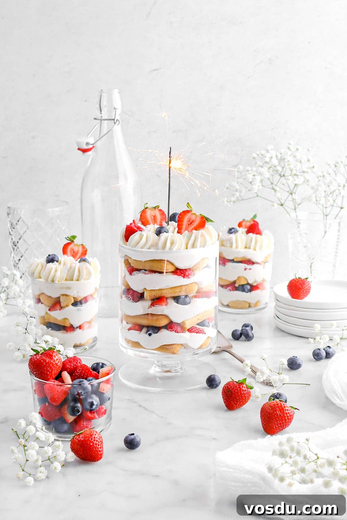 berry trifle with lit sparkler in main trifle, with two more smaller trifles behind, with white flowers and berries around.