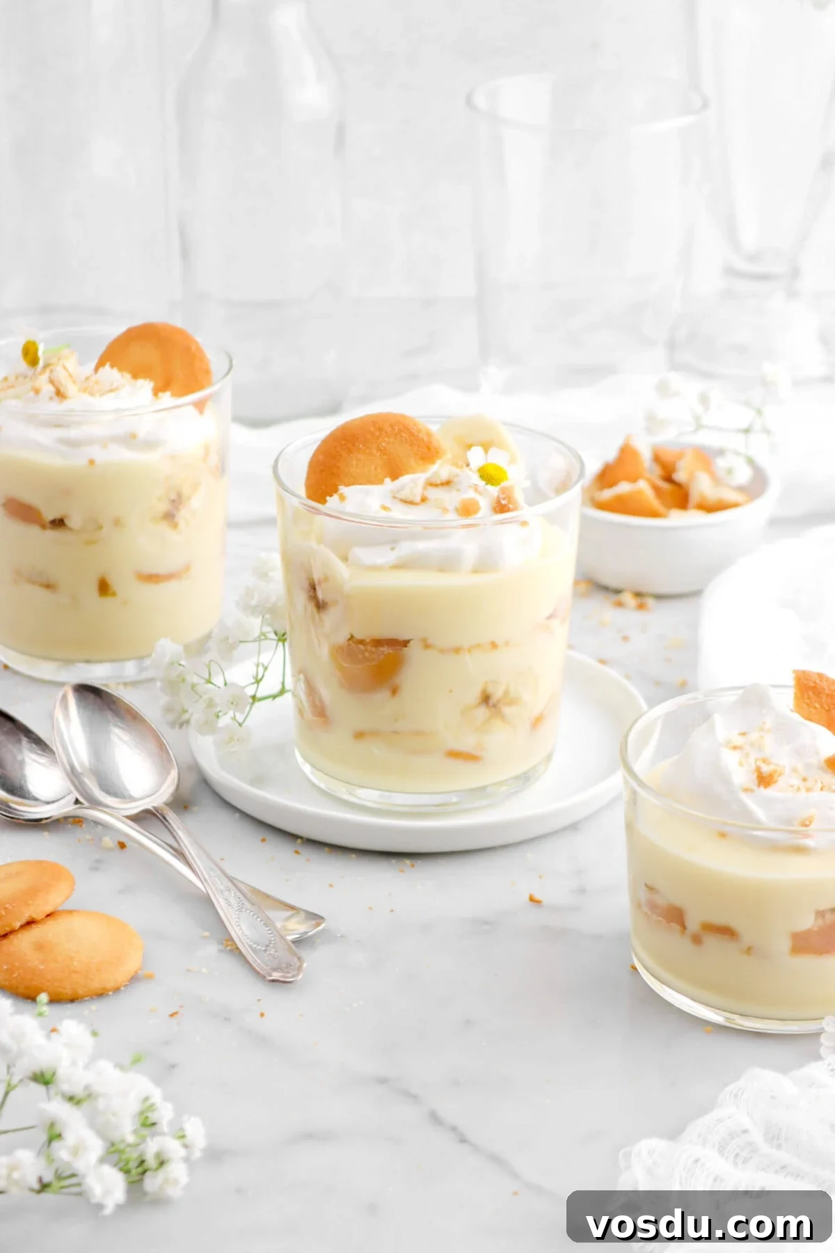glasses of banana pudding with two spoons beside, vanilla wafers and flowers around on marble surface.