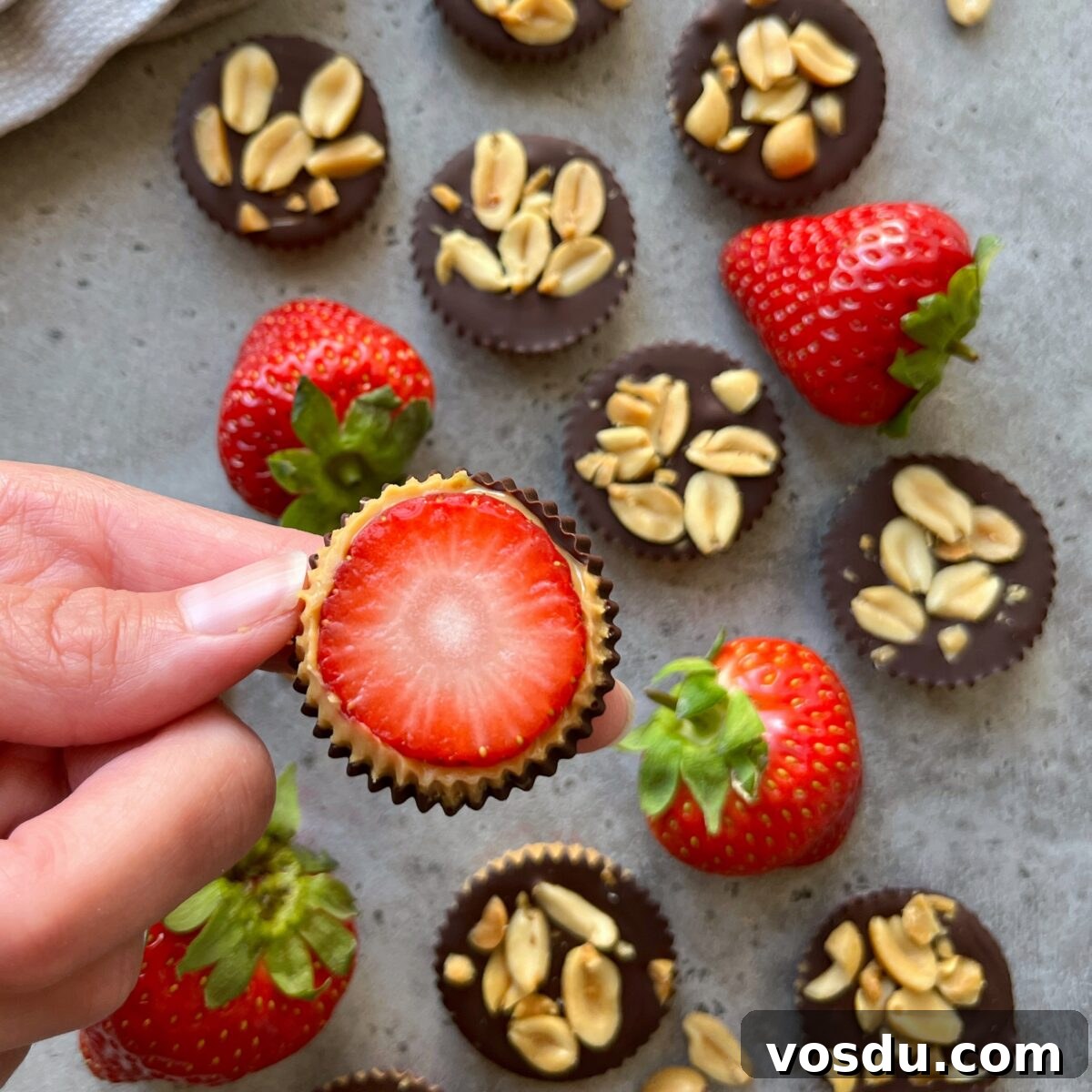 Strawberry Snickers Dessert: Creamy Peanut Caramel Bars 3 Hand holding a chocolate cup with a halved strawberry, surrounded by whole strawberries and strawberry Snickers-inspired chocolate cups topped with peanuts on a gray surface.