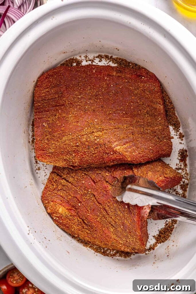 Flank steak seasoned with Greek spice blend being placed into a slow cooker with tongs