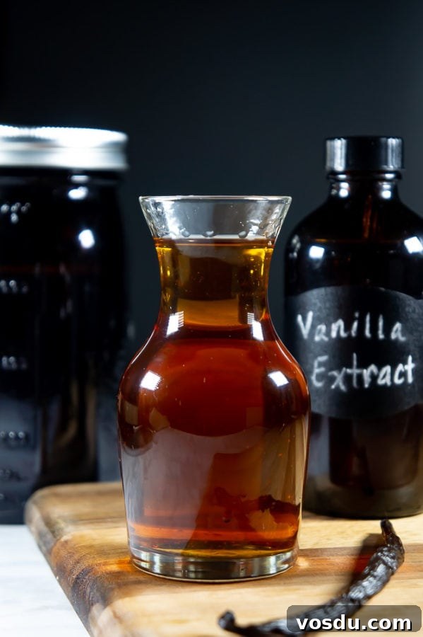 a decanter of vanilla extract on a wooden cutting board, a vanilla bean beside it, and a dark bottle of extract behind it.