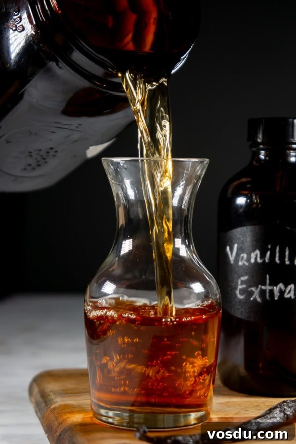 homemade vanilla extract being poured from a large amber mason jar into a decanter on a wooden cutting board beside a whole vanilla bean