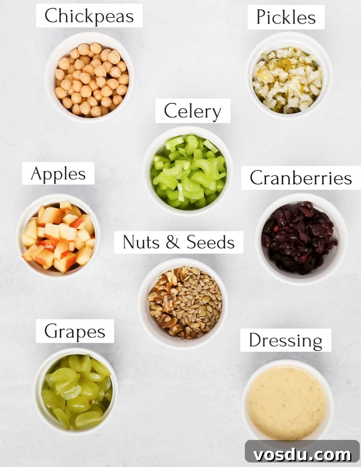 Flavorful Vegan Chickpea Salad with Lemon Herb Dressing 3 Labeled ingredients in small white bowls including: chickpeas, pickles, celery, apples, cranberries, nuts & seeds, grapes, dressing.
