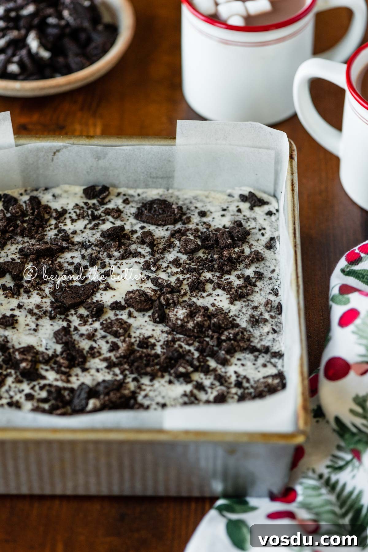 Cookies and Cream Fudge Recipe: Rich No-Bake Dessert Guide 8 Angled image of cookies and cream fudge in prepared pan with holiday themed towel, mugs of hot chocolate, and crushed chocolate sandwich cookies nearby