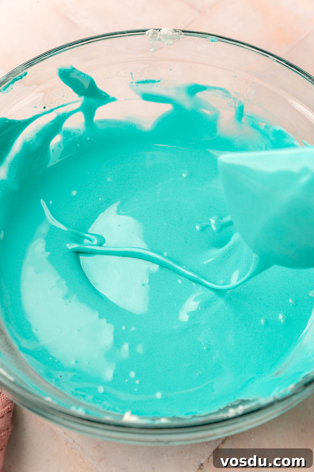 Perfect Royal Icing Recipe: Smooth, Glossy, and Pipe-Ready 8 A glass mixing bowl of teal royal icing with a spatula illustrating the ribbon test for royal icing thickness.