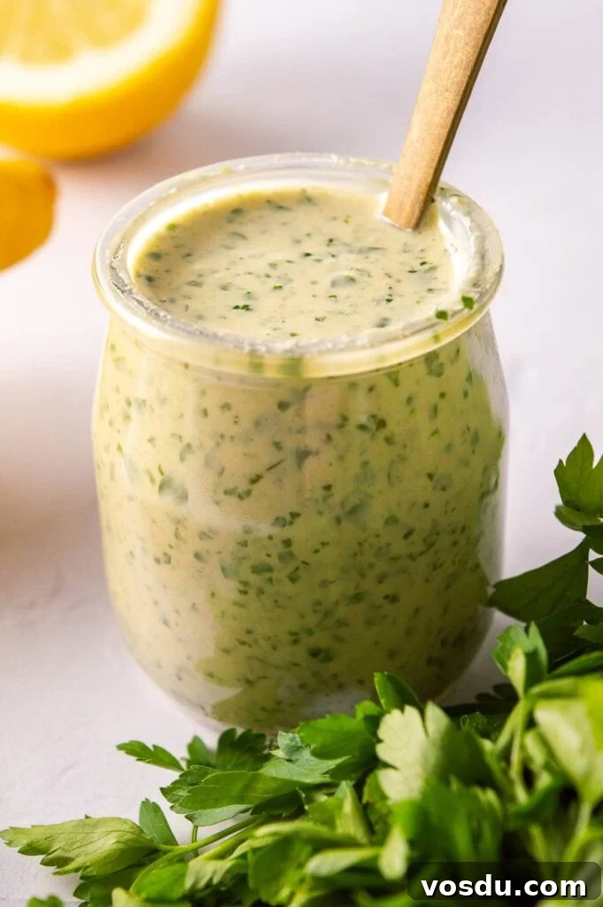A glass jar with a pale herby sauce,lemons behind it and cilantro leaves in front of it.