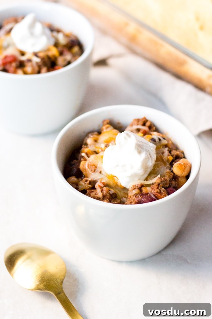 two small white bowl with gluten free beef chili in them, topped with cheese and sour cream next to cornbread and a gold spoon.