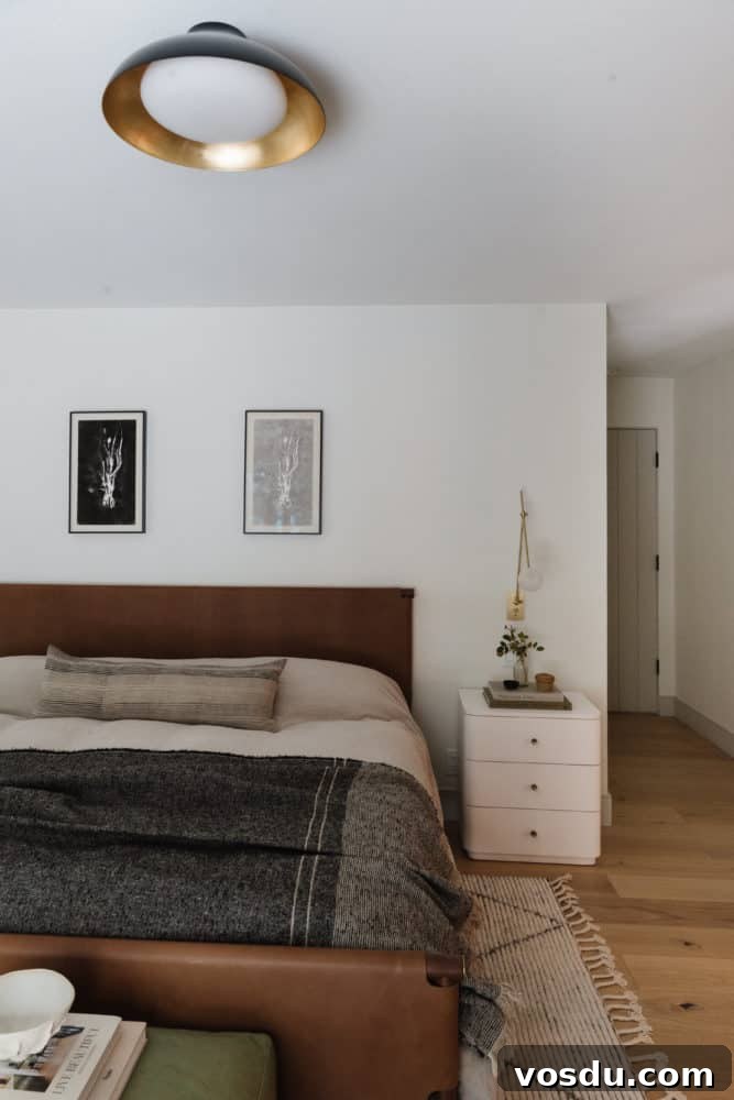 bedroom scene with a carmel eather headboard and footboard, neutral linen bedding with a charcoal throw blanket, black and white art above the headboard. White nightstand with brass and glass modern sconce next to the bed.
