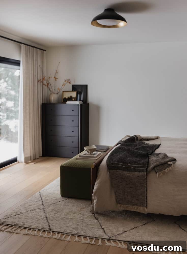 bedroom scene with medium tone wood floors, olive green velvet bench at the end of the linen duvet covered bed with a charcoal throw blanket on the bottom edge with a tall black dresser in the corner and floor to ceiling light linen curtains.