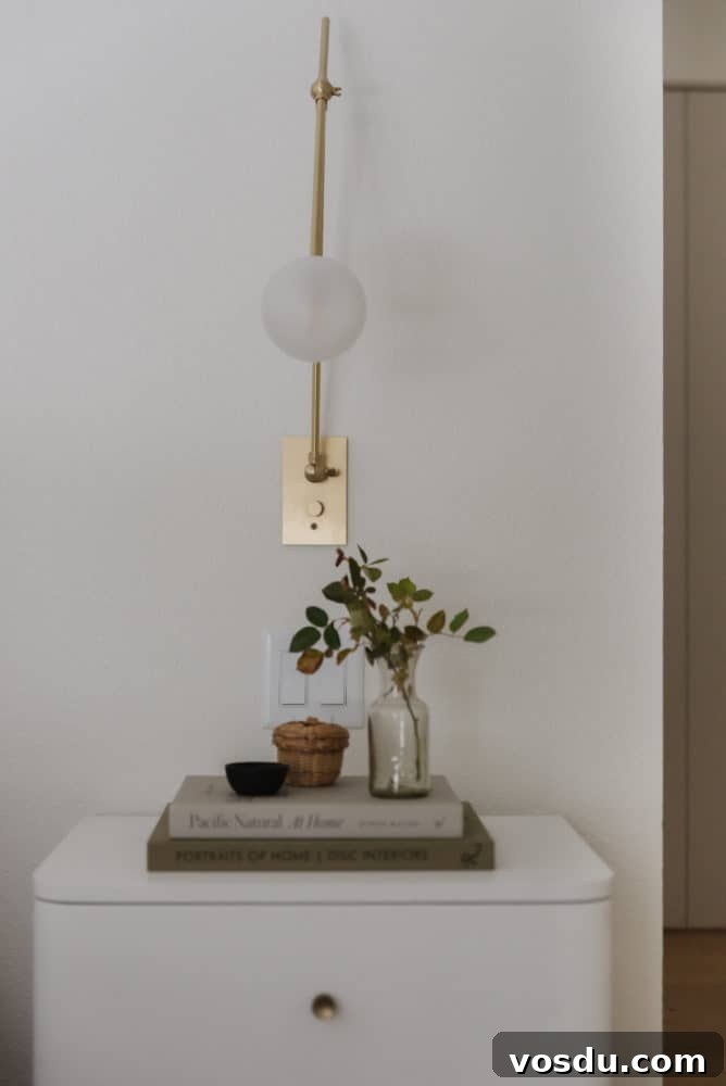 close up of white nightstand with 2 books stacked with a vase of leaves and small decorative bowls with a brass and glass modern sconce on the wall.