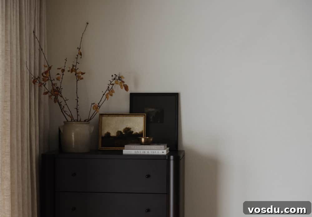 tall black dresser in a bedroom corner with linen curtain, ceramic pottery vase with tree branches inside, stacked books, and artwork.