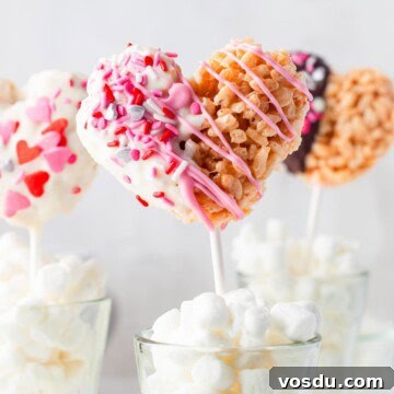 Valentine rice krispie treats hearts on sticks dipped in white and pink chocolate standing in a glass of marshmallows.