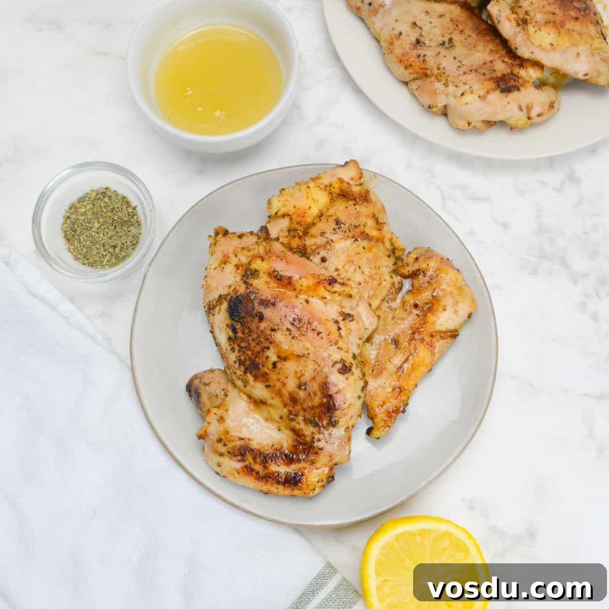 Small plate with two cooked chicken thighs. To the side sit a small bowl of honey, a bowl of oregano, a sliced lemon, a white napkin and off to the side a plate of chicken.