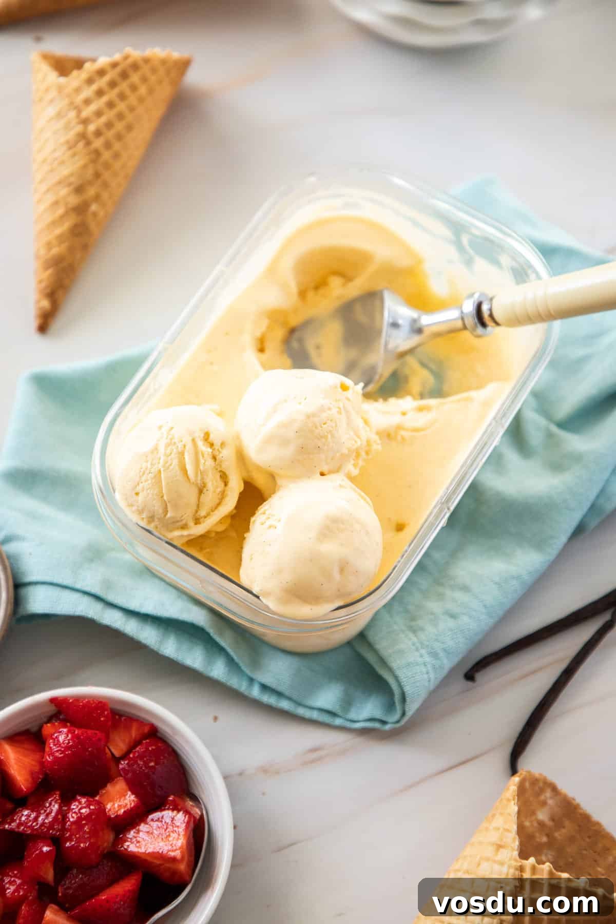 Plastic container of ice cream, with some serves scooped out, with some ice cream cones and bowl of strawberries on the edge.