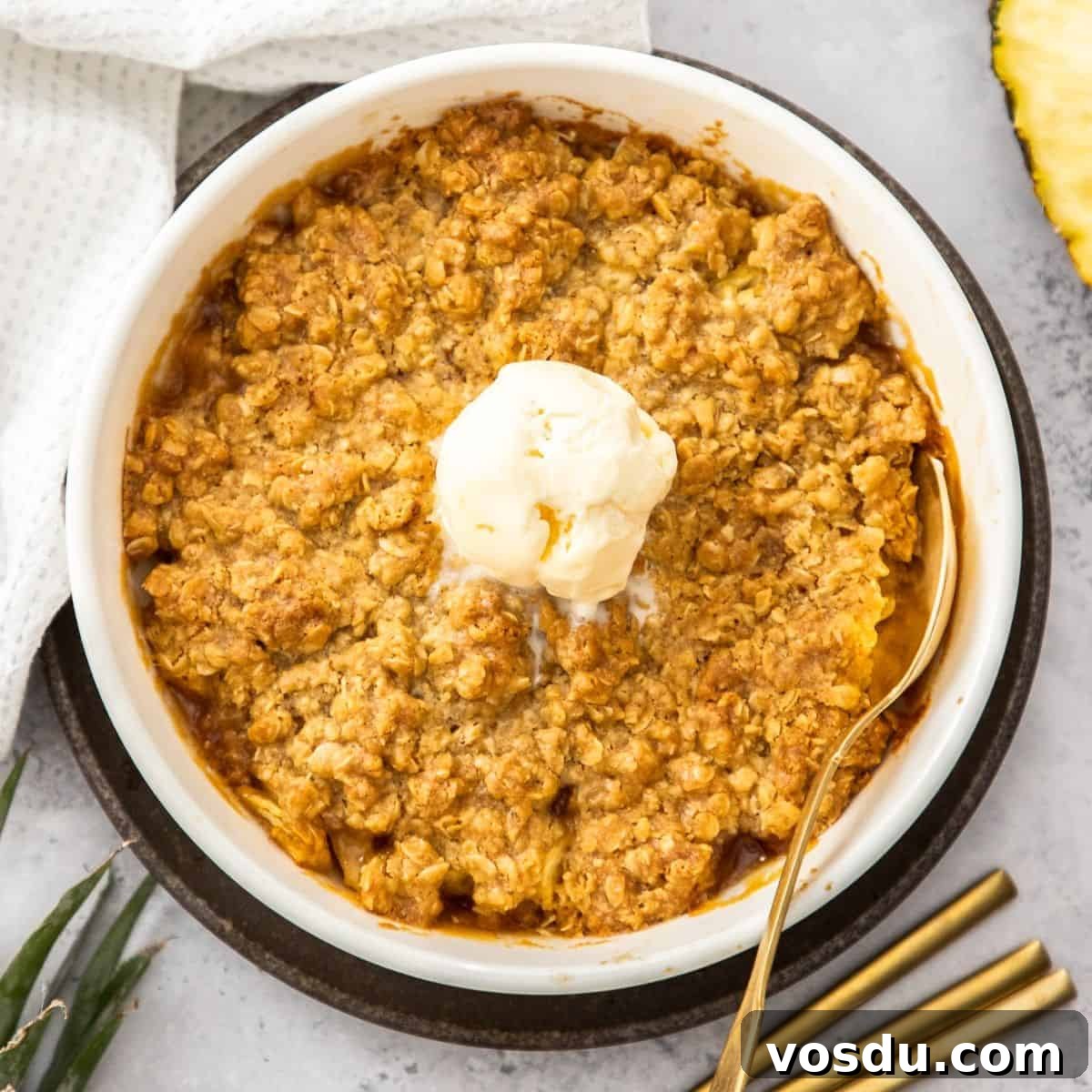 Round white dish of Pineapple Crisp, with a scoop of ice cream in the centre, and a gold spoon resting in the dish.