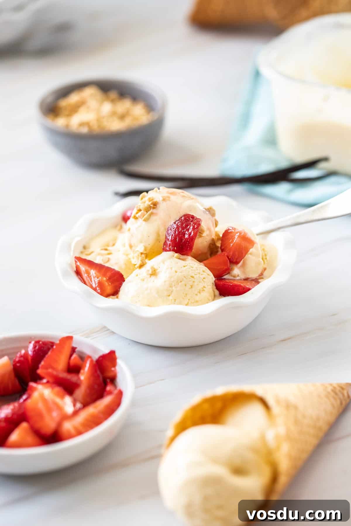 Round white bowl of ice cream topped with strawberries, with a cone with ice cream and a bowl of strawberries on the edge.