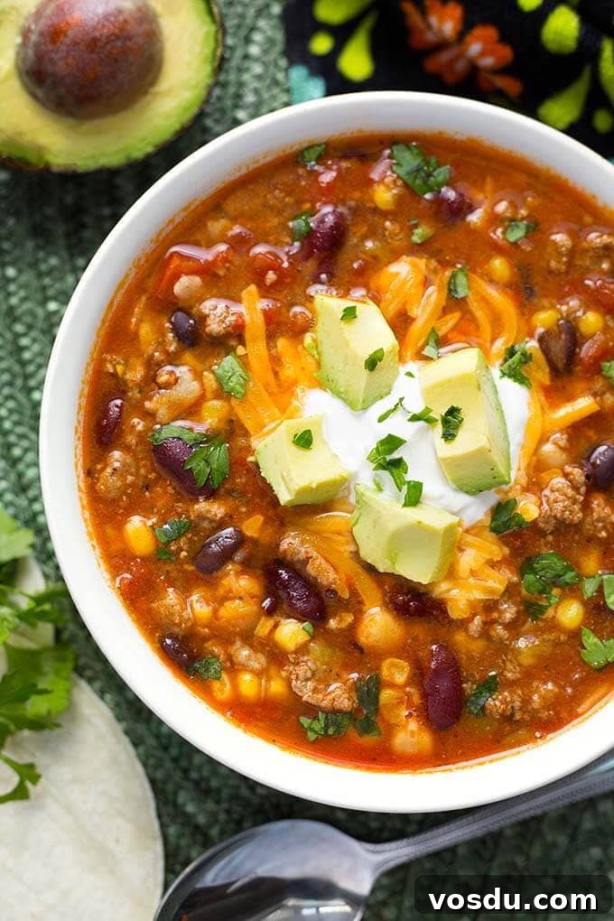 Taco Soup in a white bowl garnished with cubed avocados next to spoon all on green mat