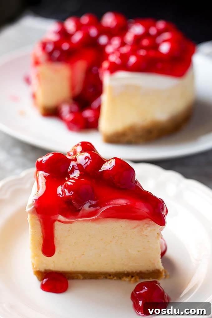 Slice of New York Cheesecake topped with canned cherries on white plate in front of rest of cheesecake