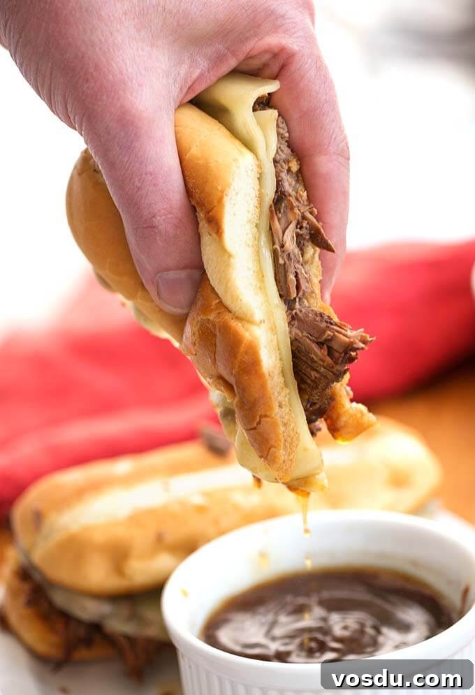 Hand dipping French Dip Sandwich in small white bowl of brown sauce
