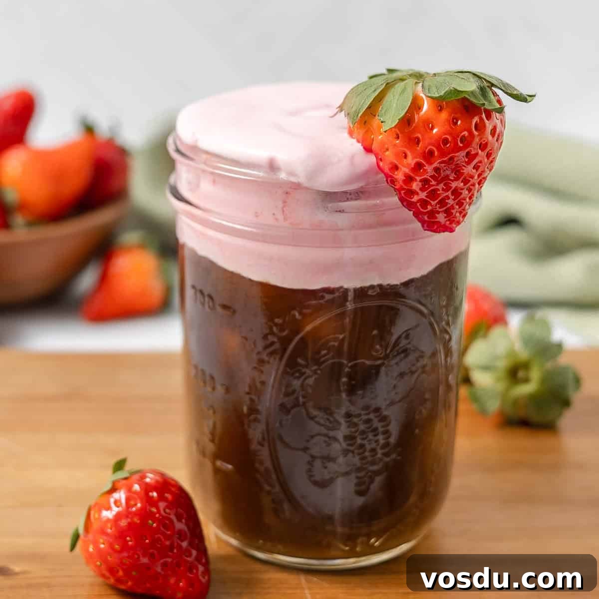 A glass jar filled with iced coffee topped with pink foam and garnished with a fresh strawberry, with more strawberries in the background.