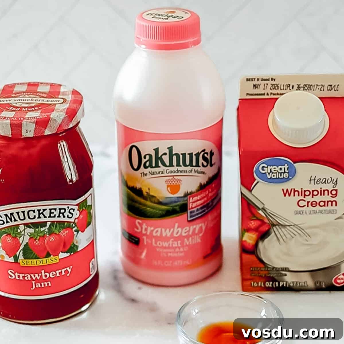 A jar of strawberry jam, a bottle of strawberry low-fat milk, and a carton of heavy whipping cream are placed on a countertop.